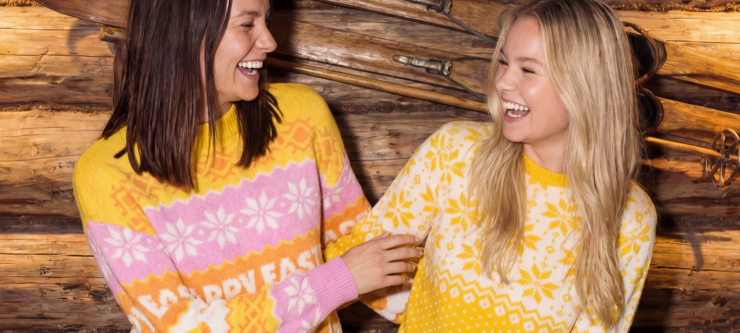 Two women laughing in pink and yellow Fair Isle sweaters against a wooden cabin wall with old skis