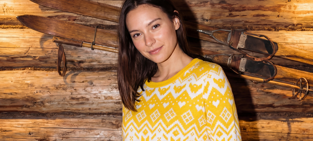 Smiling woman in a yellow patterned sweater standing against a wooden wall with vintage skis